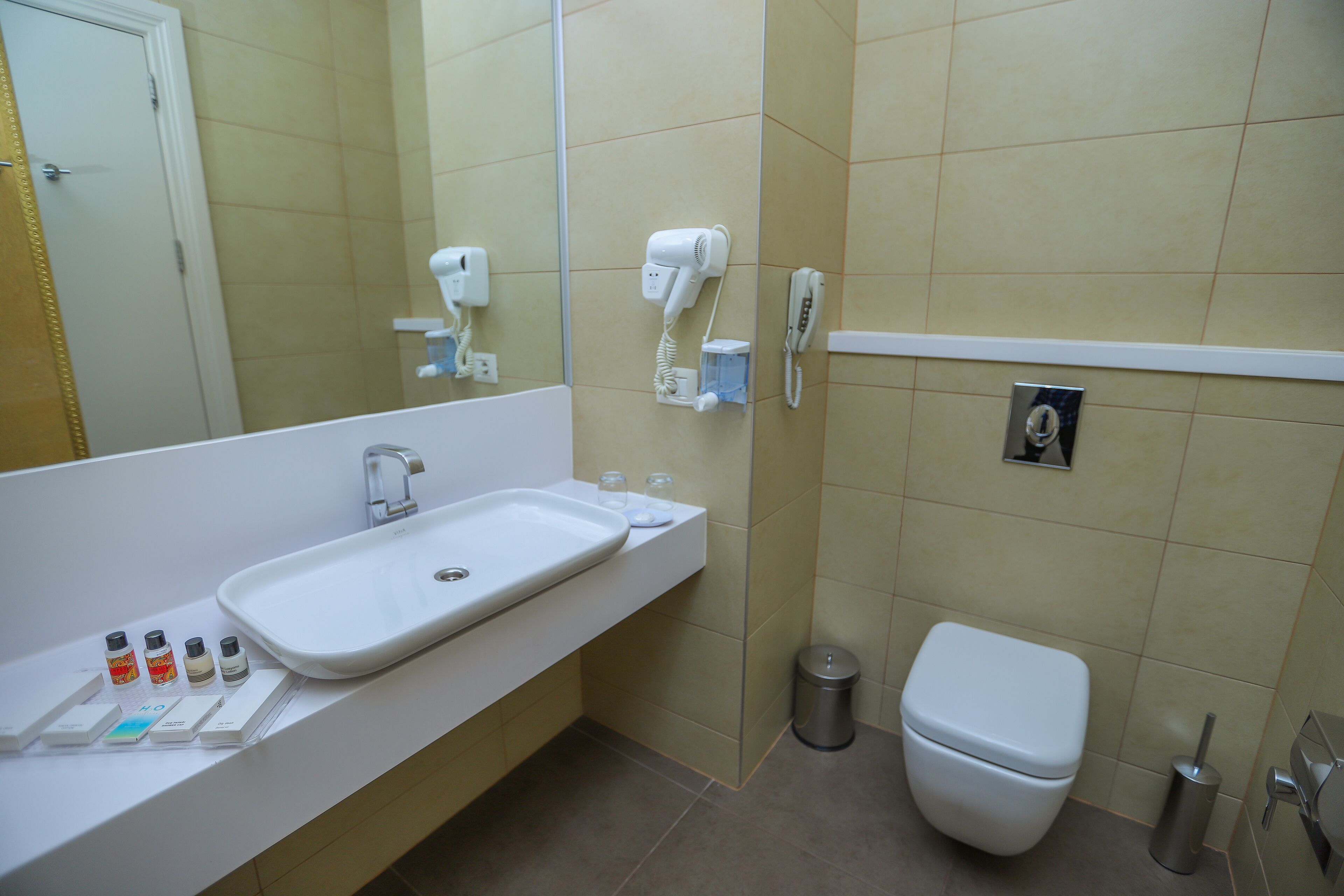 Une salle de bain équipée d’un lavabo, d’un miroir, d’un sèche-cheveux, d’un téléphone, de toilettes et d’une poubelle.