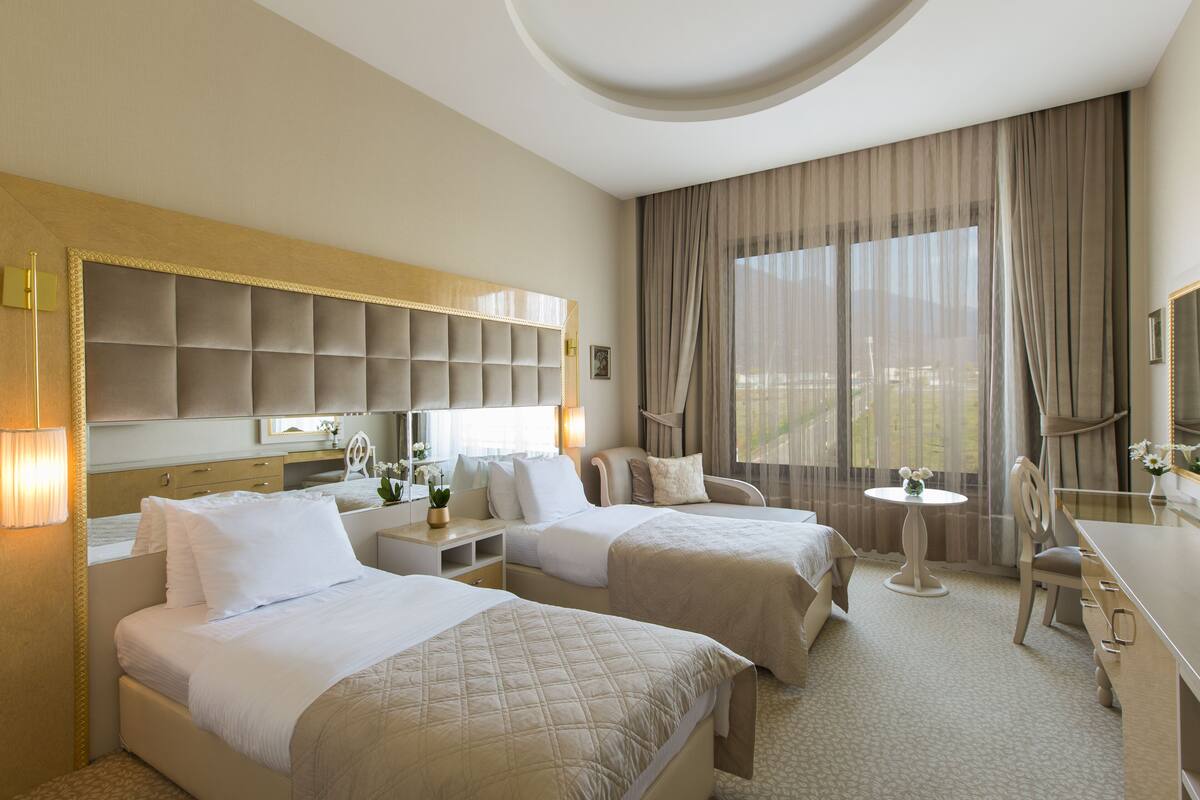 A hotel room with two beds, a desk, a chair, a small table, and a large window with curtains.