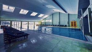 Una piscina interior con paredes de vidrio transparente, un techo iluminado por el sol y una fila de tumbonas a un lado.