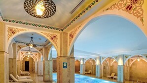 Una piscina con un diseño geométrico de mosaicos, rodeada de sillones de descanso y arcos con detalles ornamentales.
