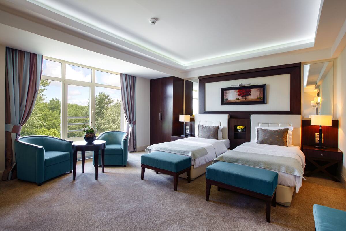 Habitación de hotel con dos camas, una zona de estar con mesa y sillas, ventanas amplias con cortinas y un cuadro en la pared.