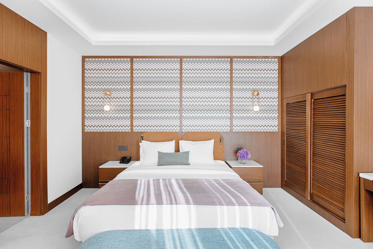 A bedroom with a large bed, wooden headboard, nightstands, a wooden wardrobe, and a decorative wall panel.