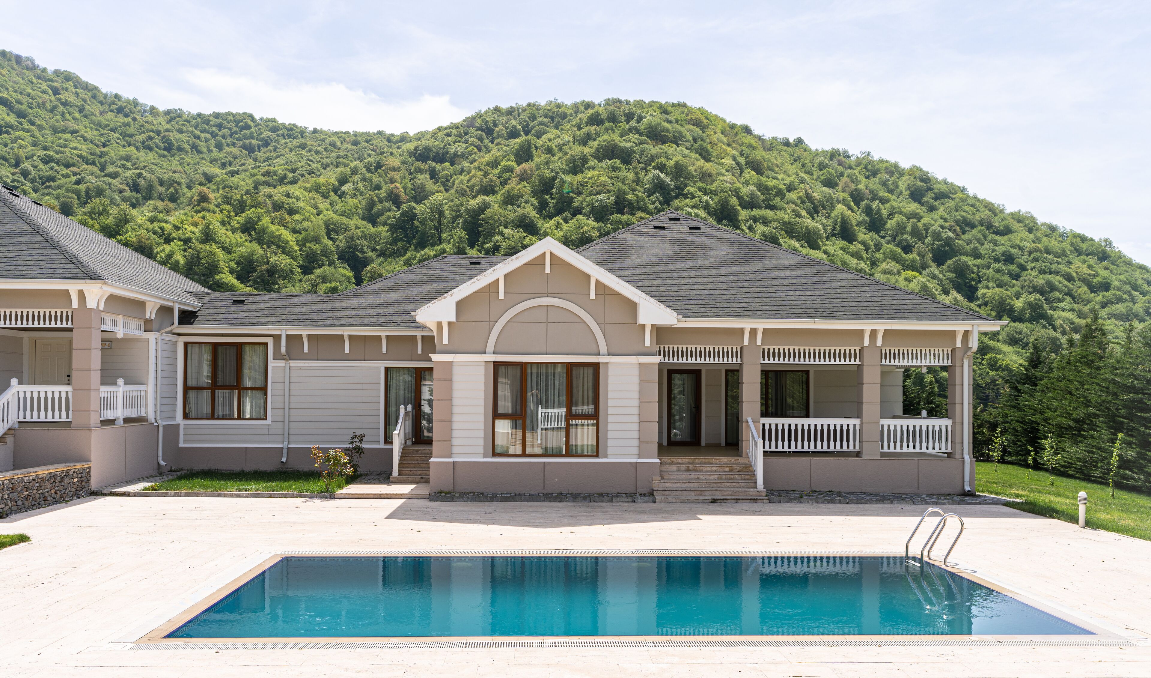 A two-story house with a swimming pool in front, surrounded by a forested hillside.