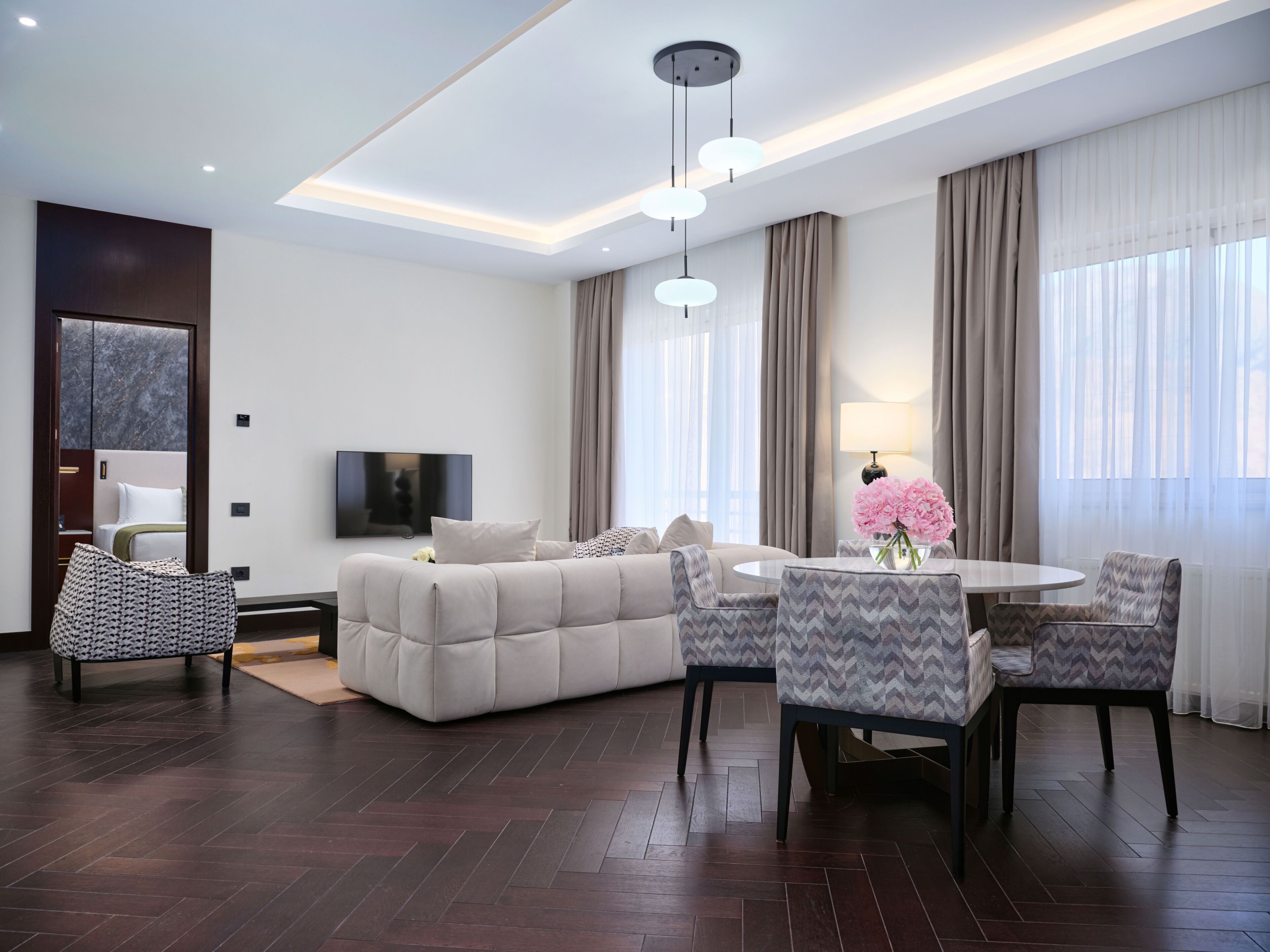 A modern living room with a sofa, dining table, chairs, a TV, and a large window with curtains.