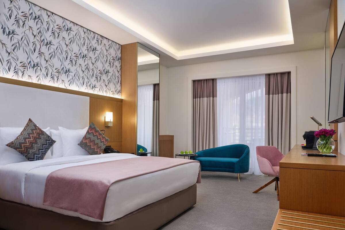 A modern hotel room with a large bed, a desk, a blue armchair, and a wall-mounted TV.
