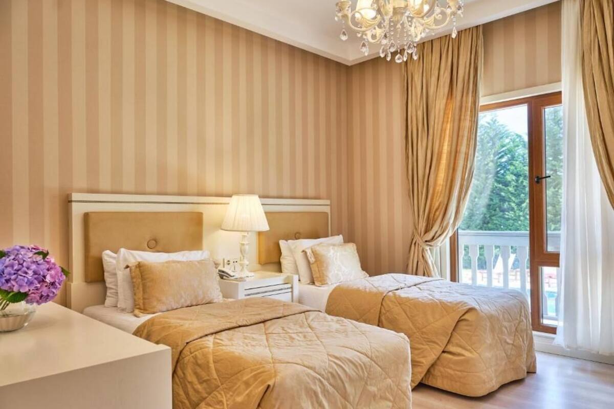 A hotel room with two beds, a chandelier, a balcony with curtains, and a view of greenery.