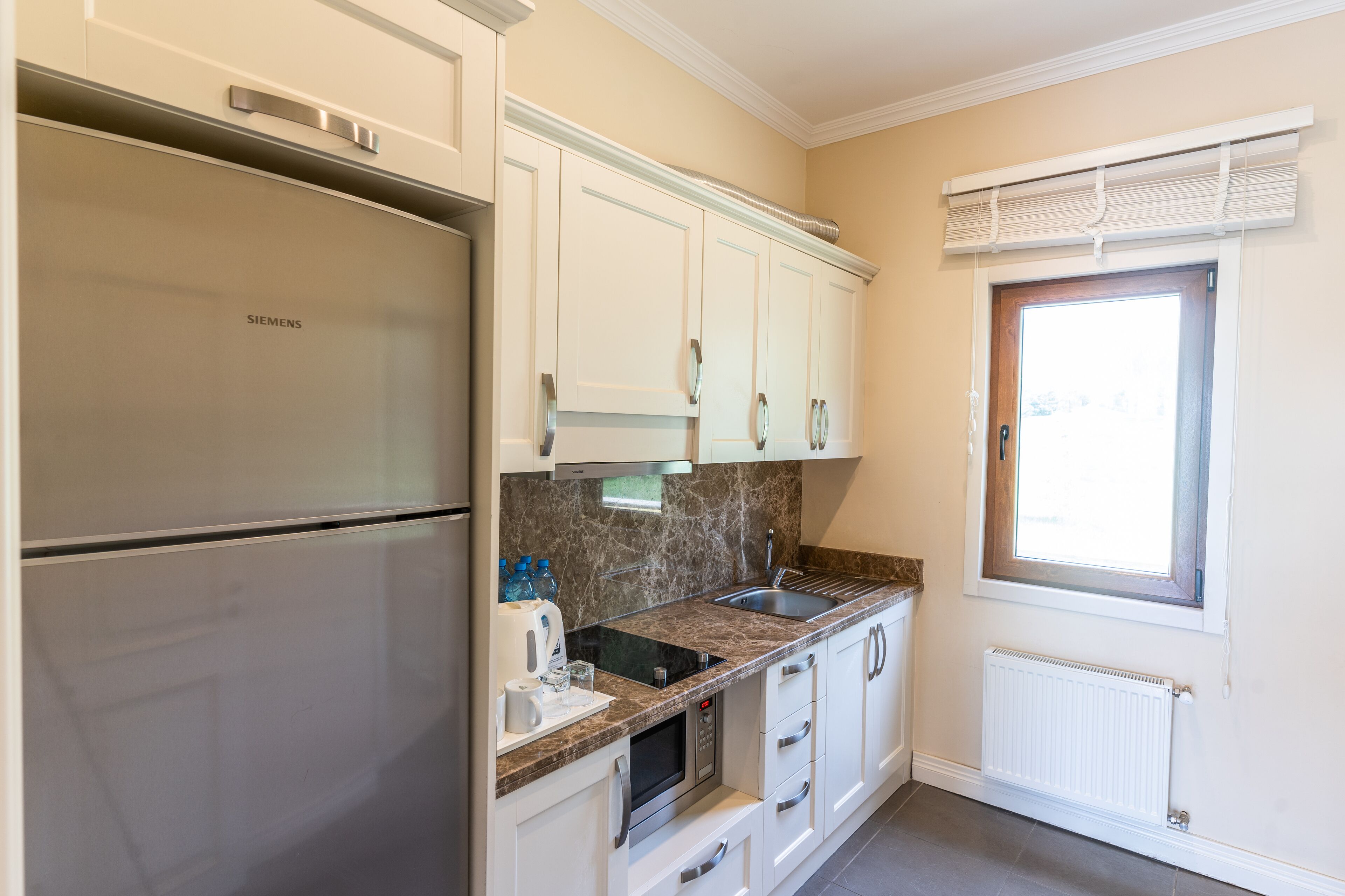 A modern kitchen with white cabinets, a stainless steel refrigerator, a built-in oven, and a window with blinds.