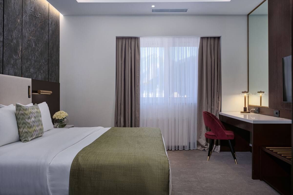 A hotel room with a bed, a desk, a chair, a mirror, and a window with curtains.