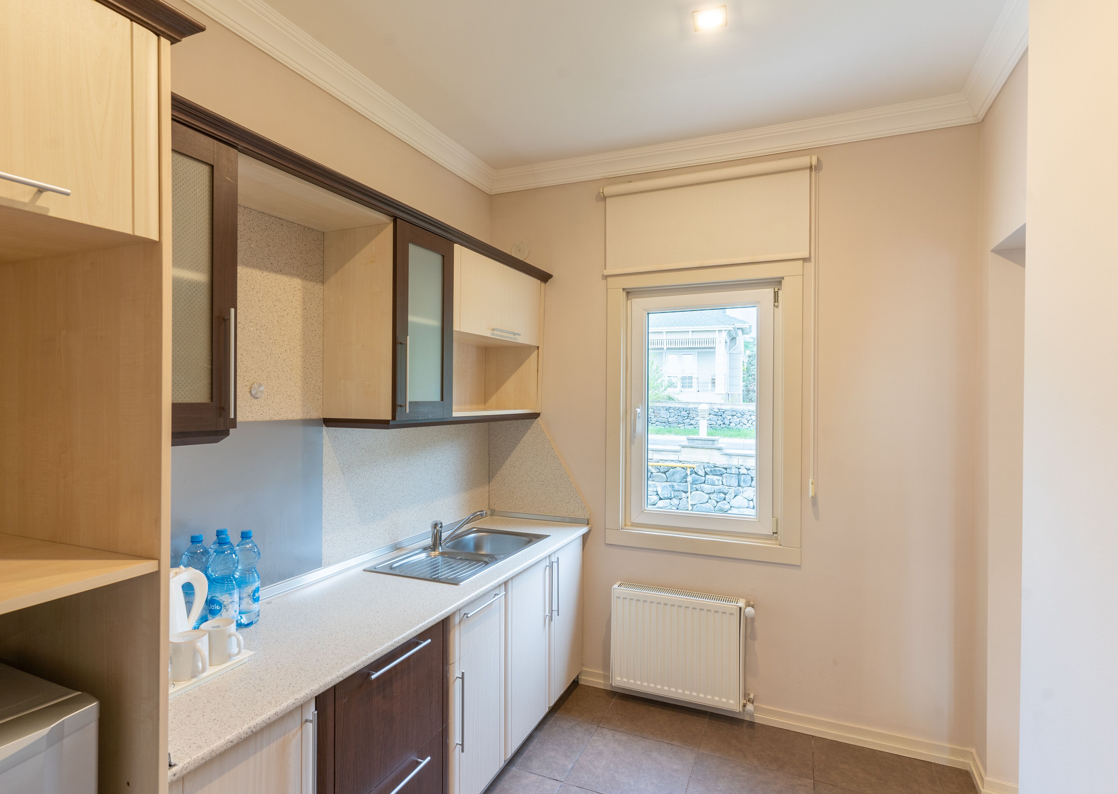 A compact kitchen with a built-in sink, a window with a view of a stone wall and a house, and a radiator.