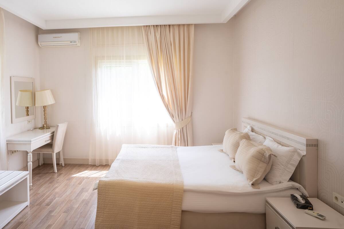 A hotel room with a bed, bedside table, lamp, desk, chair, and an air conditioning unit.