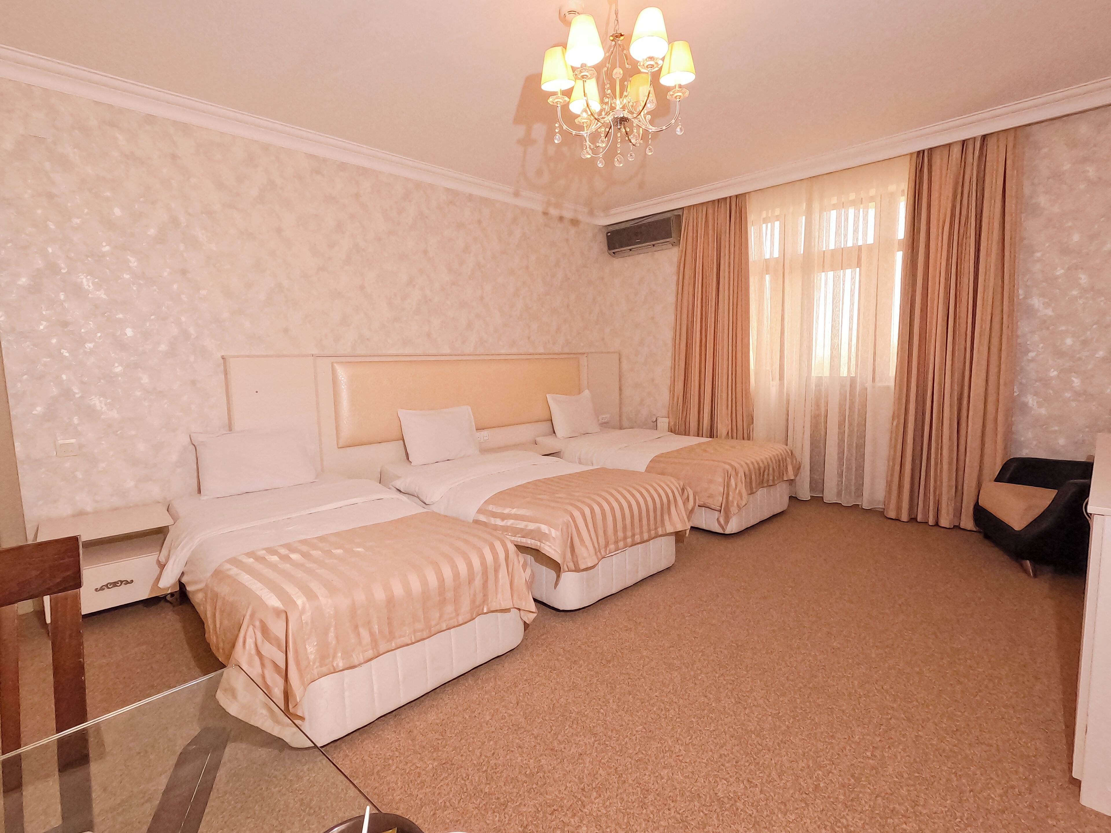 A hotel room with two beds, a chandelier, a large window with curtains, and a wooden chair.