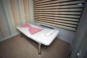 Habitación con una cama individual, piso de madera, ventana con persianas horizontales y mesita de noche con una almohada y una toalla rosa.