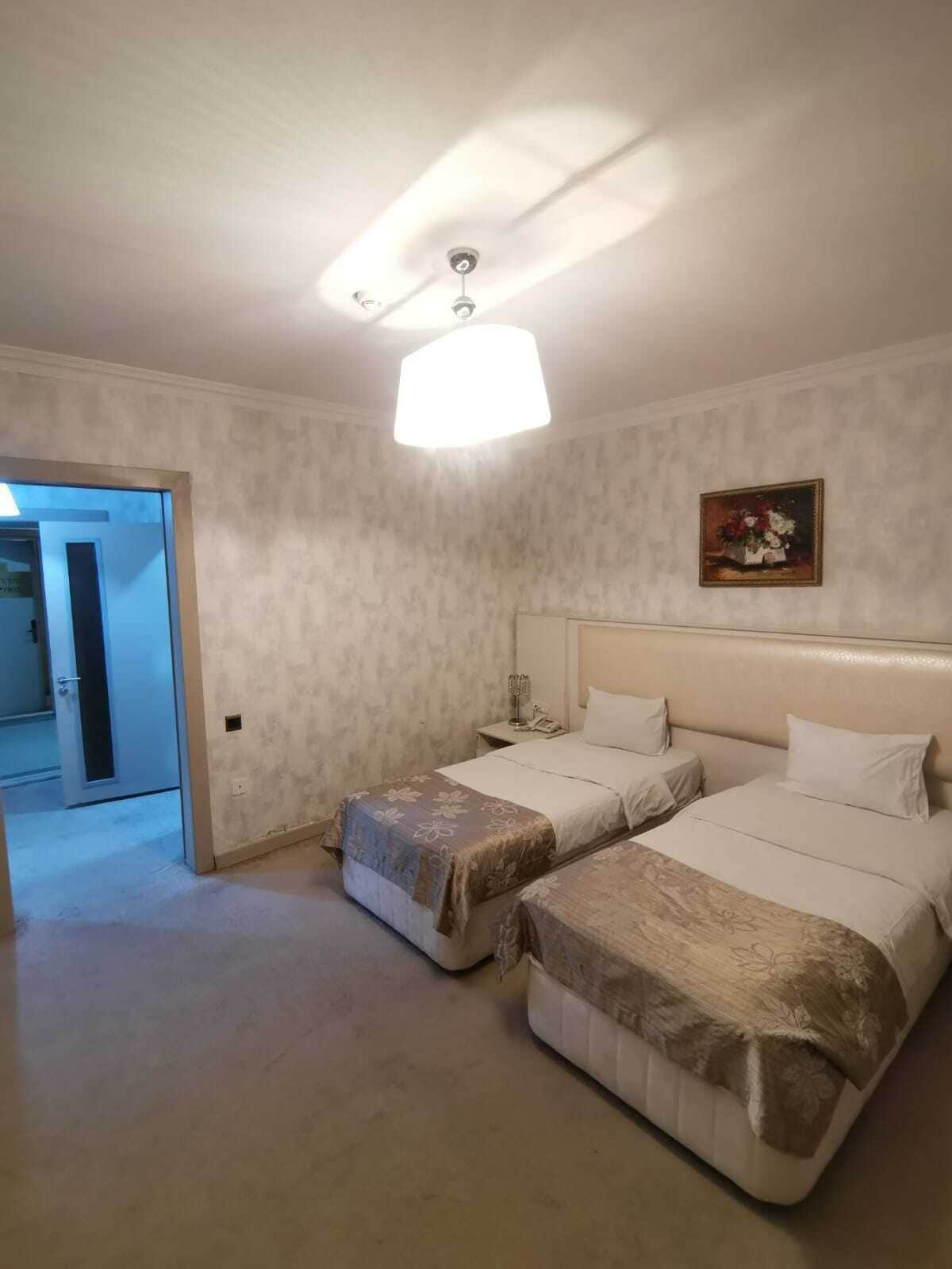 A hotel room with two beds, a ceiling light, a painting on the wall, and a door leading to another room.