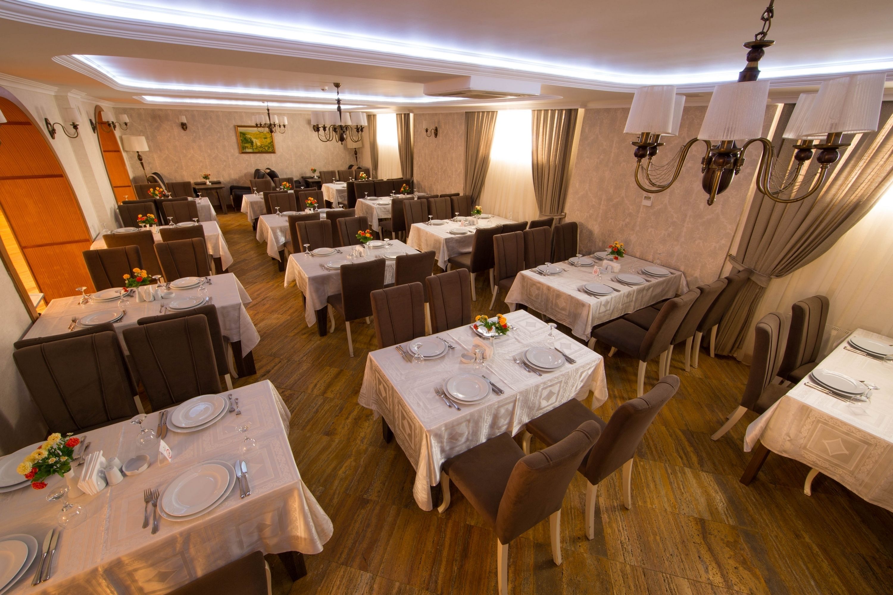A dining area with multiple tables set for service, featuring white tablecloths, brown chairs, and chandeliers.