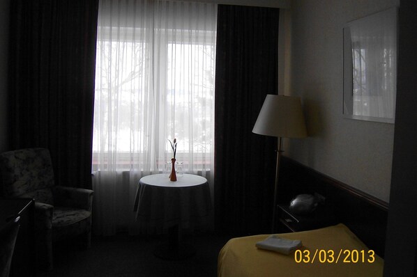 A hotel room with a bed, a chair, a small round table with a vase, and a lamp.