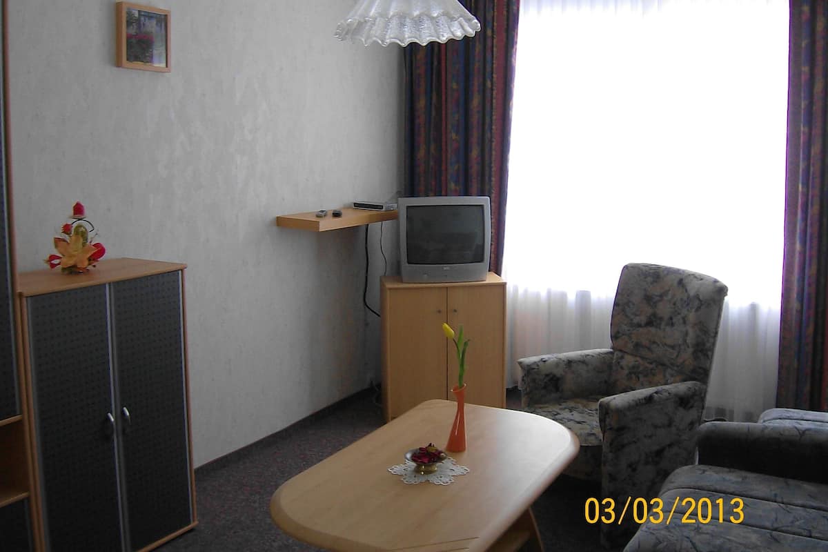 A small living room with a television on a wooden stand, a single armchair, a small table with a vase of flowers, and a cabinet.