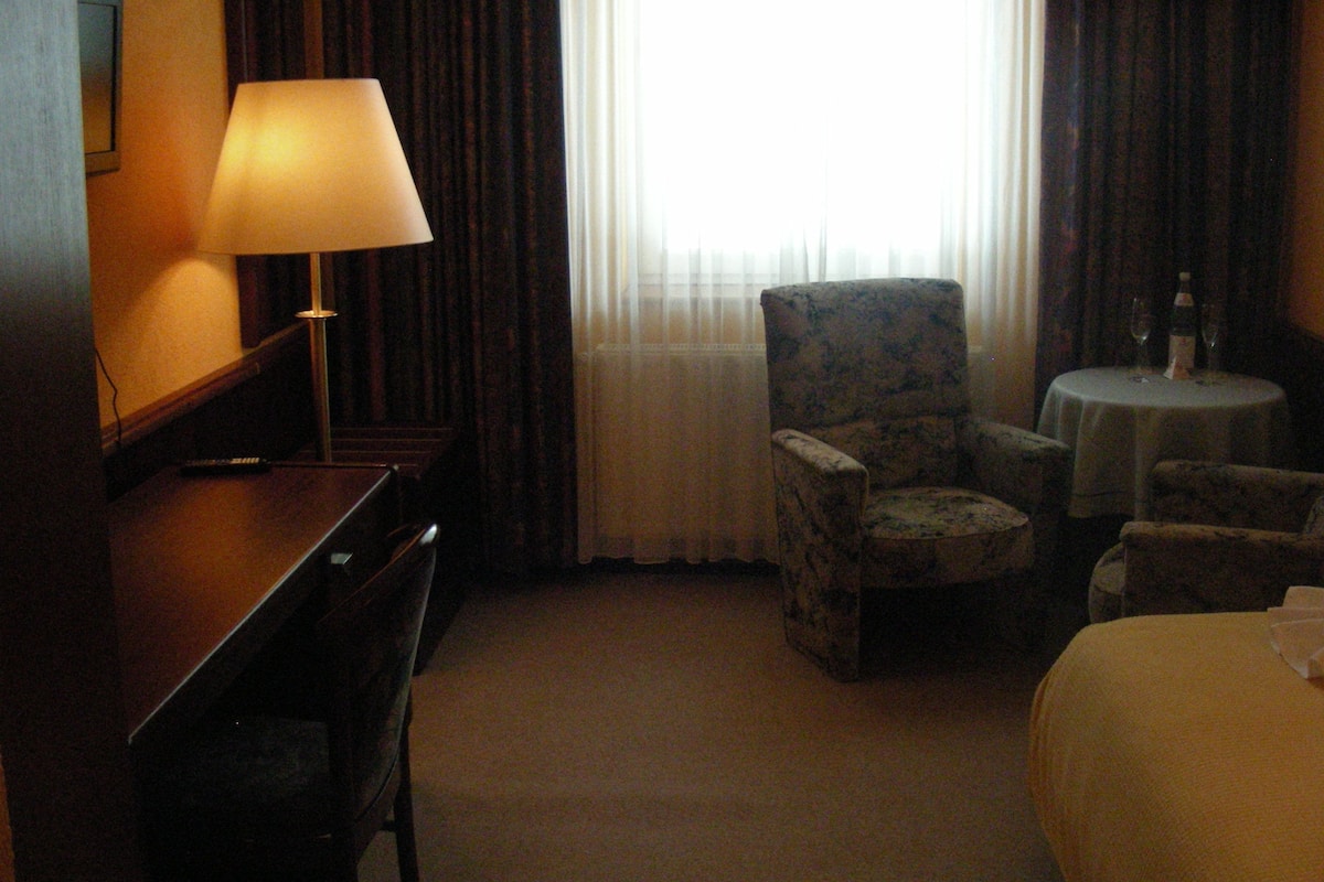 A hotel room with a desk, a chair, a bed, a round table with a bottle and glasses, and a window with curtains.