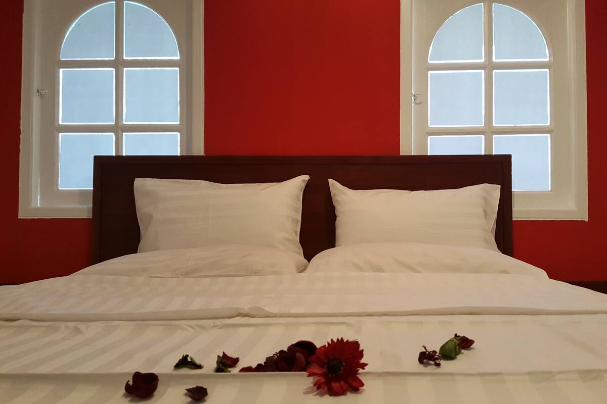 A bed with white linens, two windows with arched tops, and red walls.