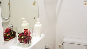 Casa de banho com lavatório branco, sanita e chuveiro. Existem duas prateleiras com produtos de higiene pessoal e um espelho.