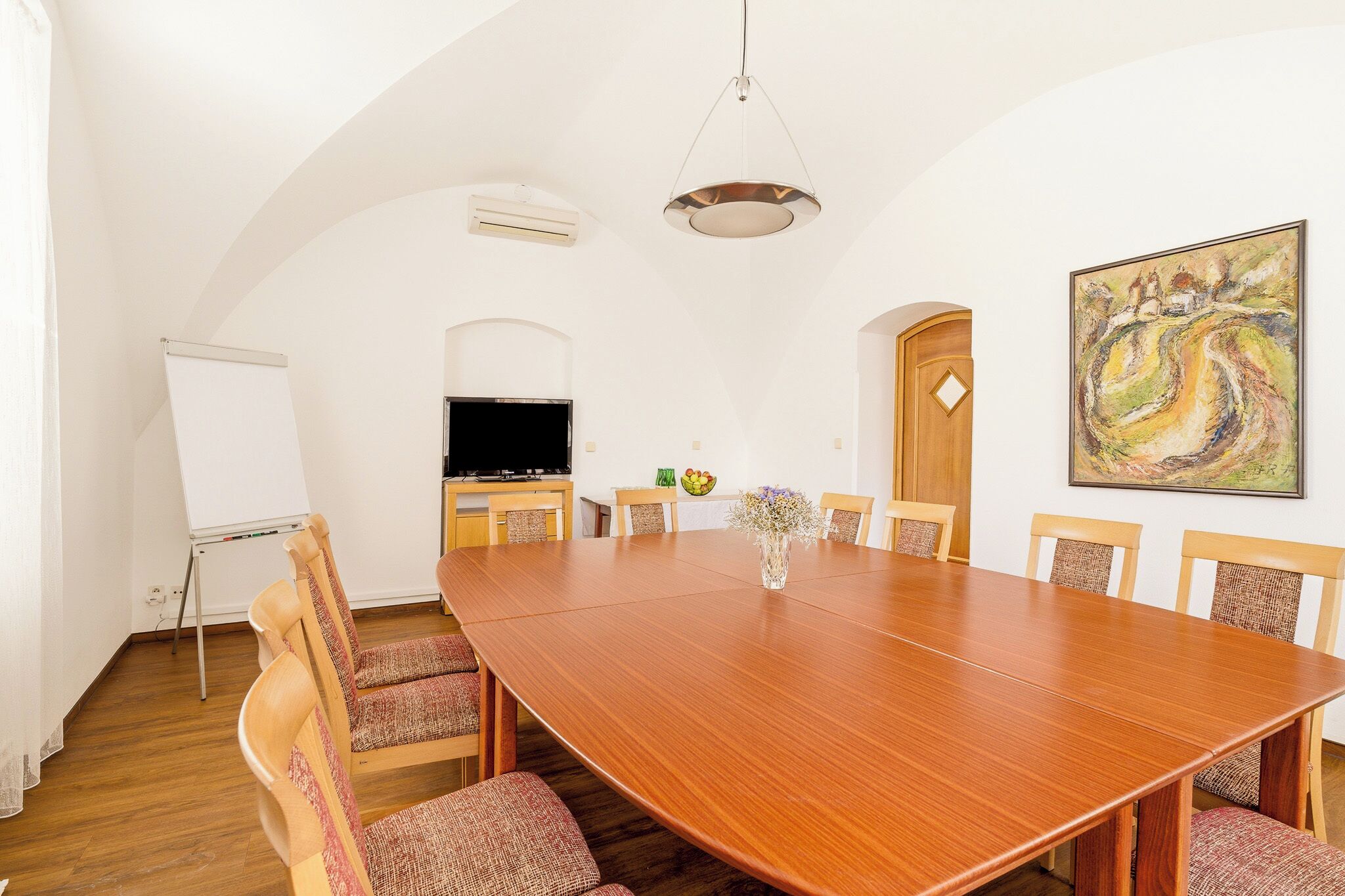 A conference room with a large wooden table, chairs, a whiteboard, a television, and a painting on the wall.