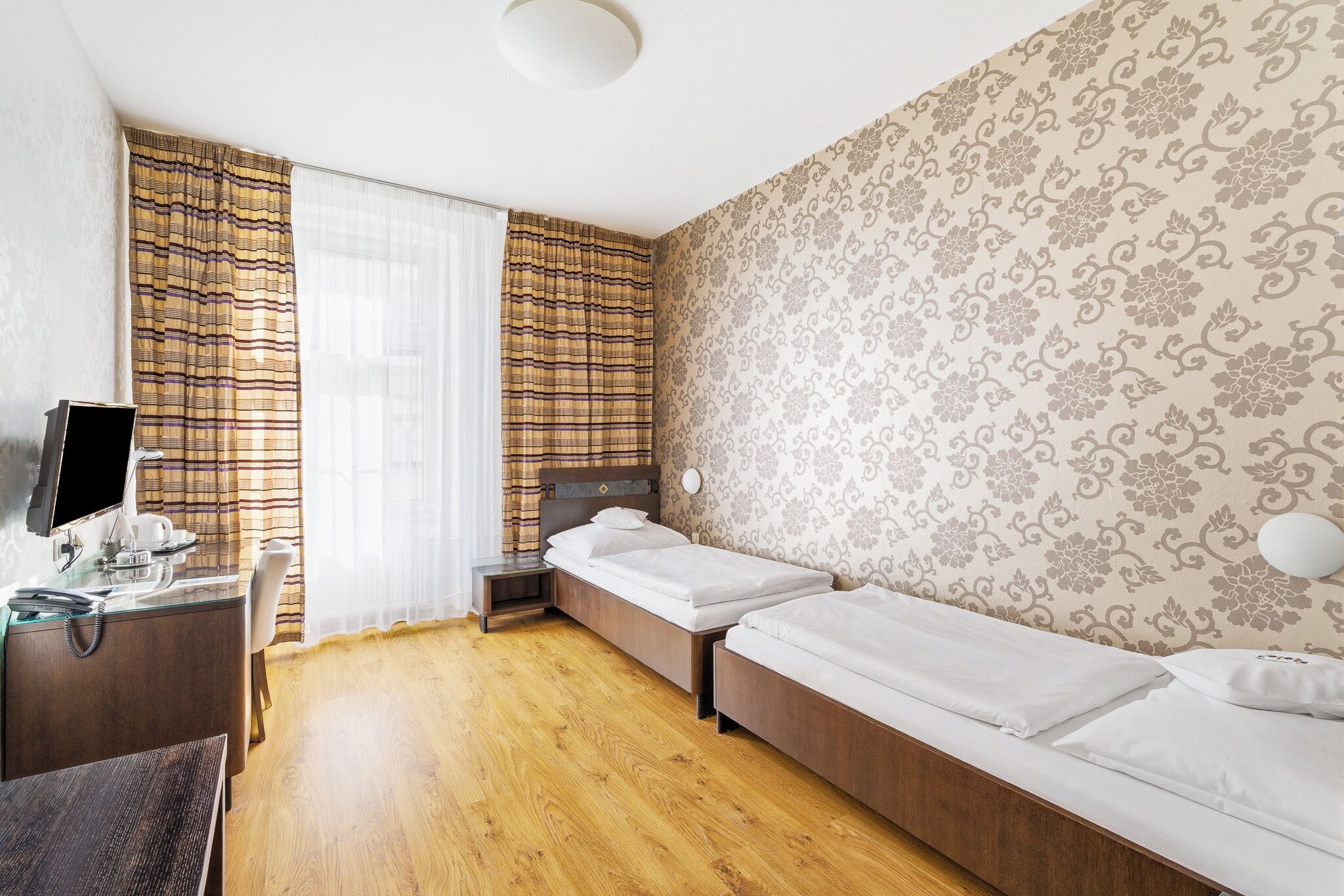 A hotel room with a wooden floor, two single beds, a desk with a computer, and a TV.