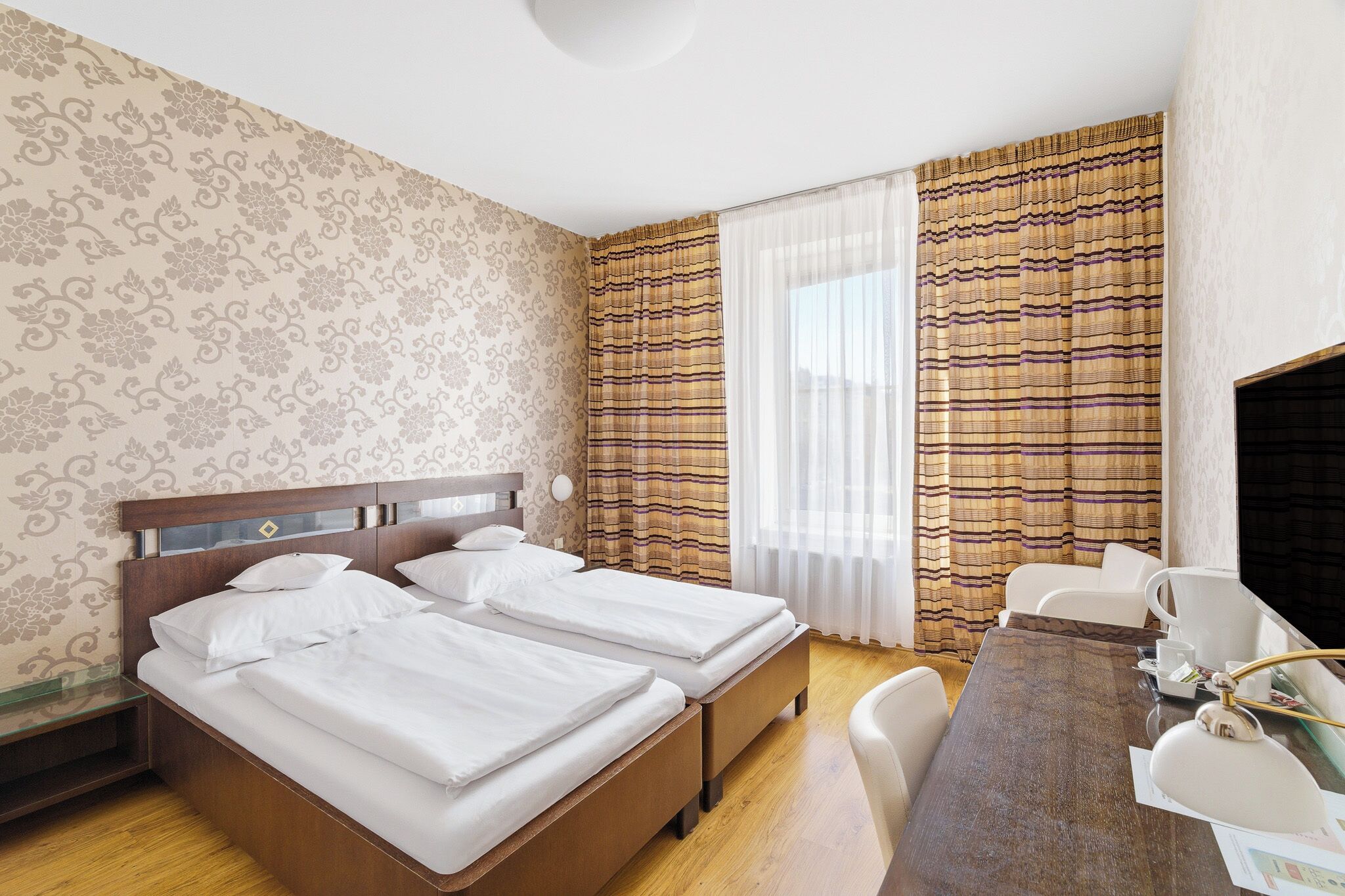 Una camera d'albergo con due letti, una testiera in legno, una scrivania con sedie e un'ampia finestra con tende.
