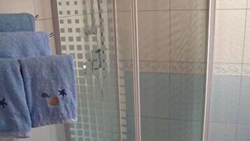 Ducha con cerramiento de vidrio, pared con azulejos blancos, un cubo de metal y dos toallas colgadas.