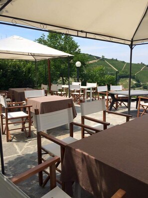 An outdoor dining area with tables and chairs under a canopy, set against a backdrop of green hills. - La Terrazza del Barbaresco (Neviglie)