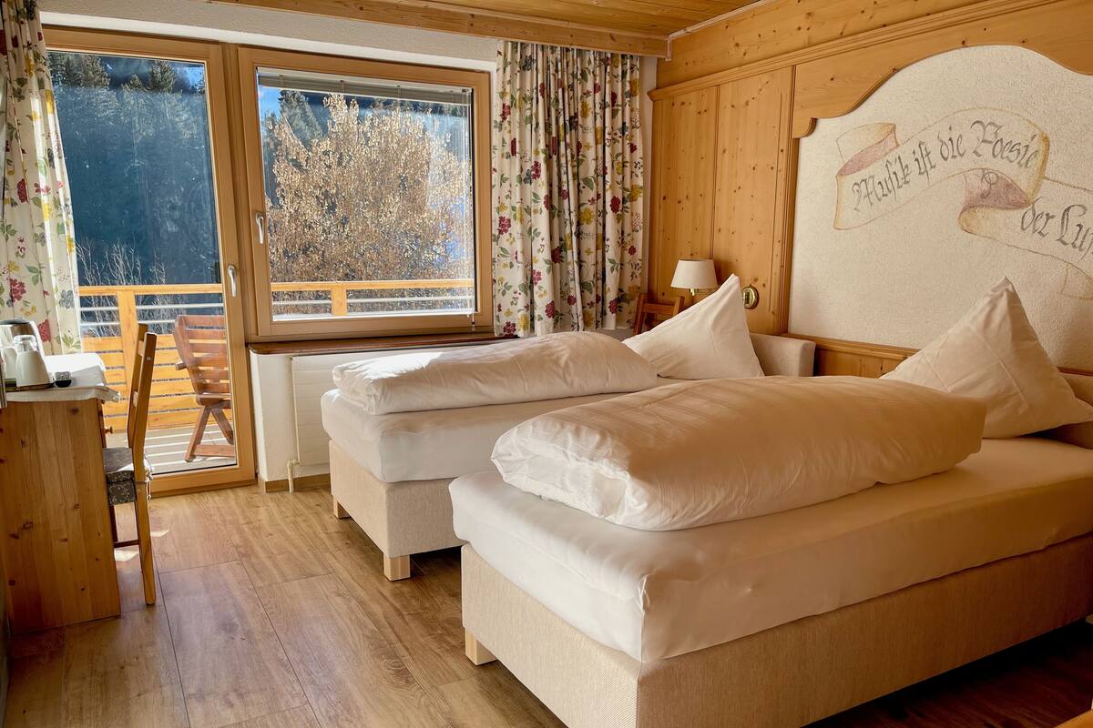 A cozy bedroom with two beds, wooden flooring, a window with curtains, and a wooden headboard with a decorative plaque.