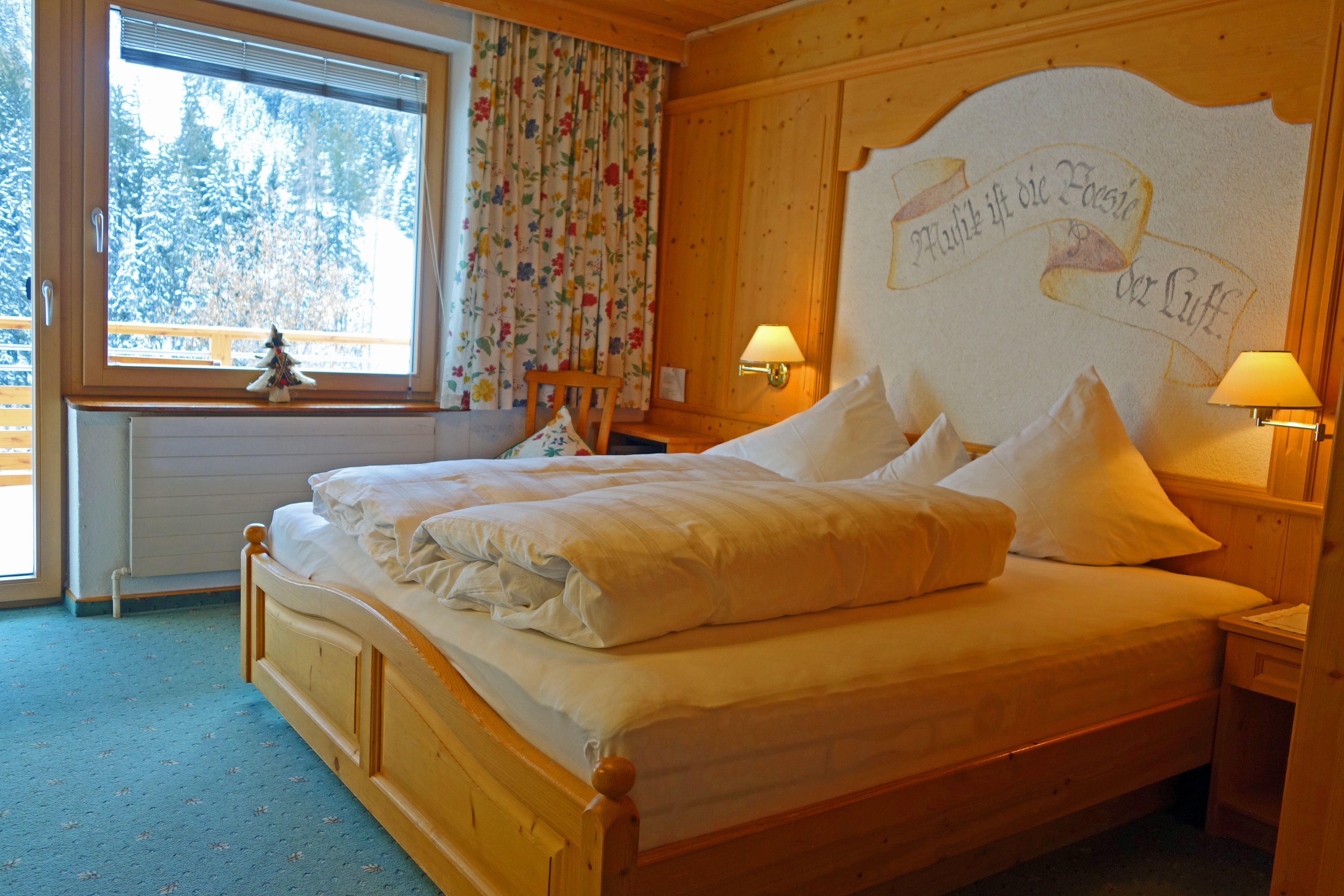 A wooden bed with white bedding, a nightstand with a lamp, a window with a view of trees, and a wall with a decorative banner.