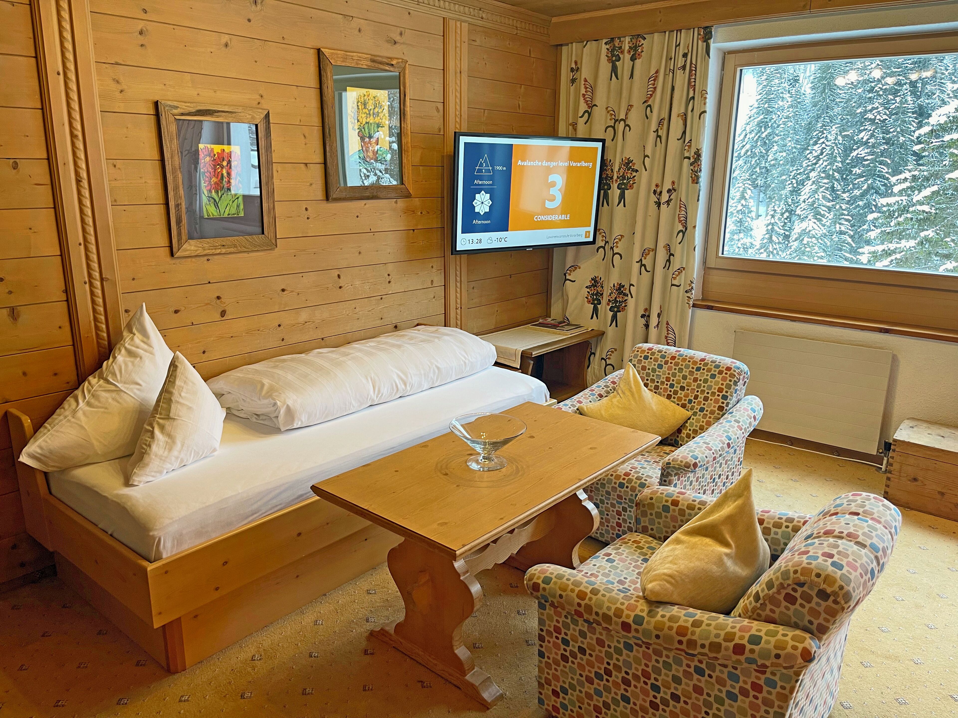 Ein gemütliches Zimmer mit einem hölzernen Bett, einem kleinen Tisch, einem farbenfrohen Sofa und einem Fernseher, der die Lawinengefahr anzeigt.