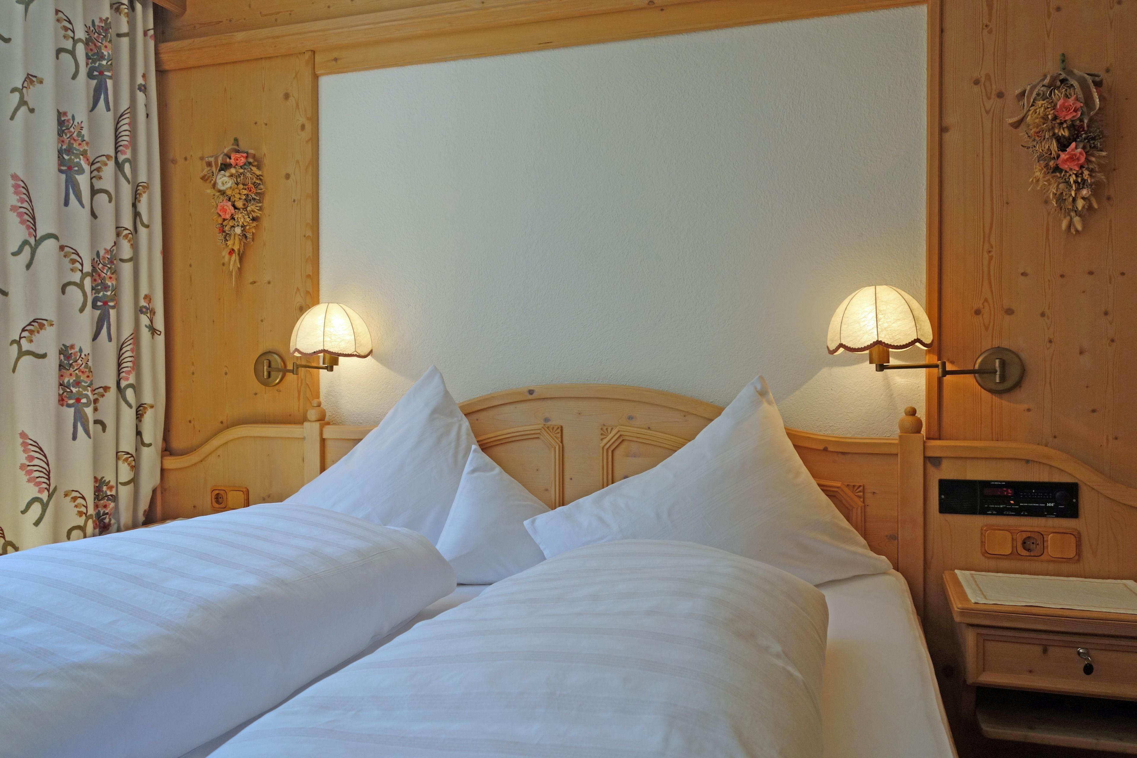 Una cama doble con sábanas blancas, dos lámparas fijadas a la pared, un cabecero de madera y una mesita de noche con un radio.