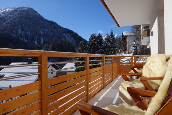 A balcony with wooden railings, a fur-covered chair, and a view of snow-covered houses and a mountain.