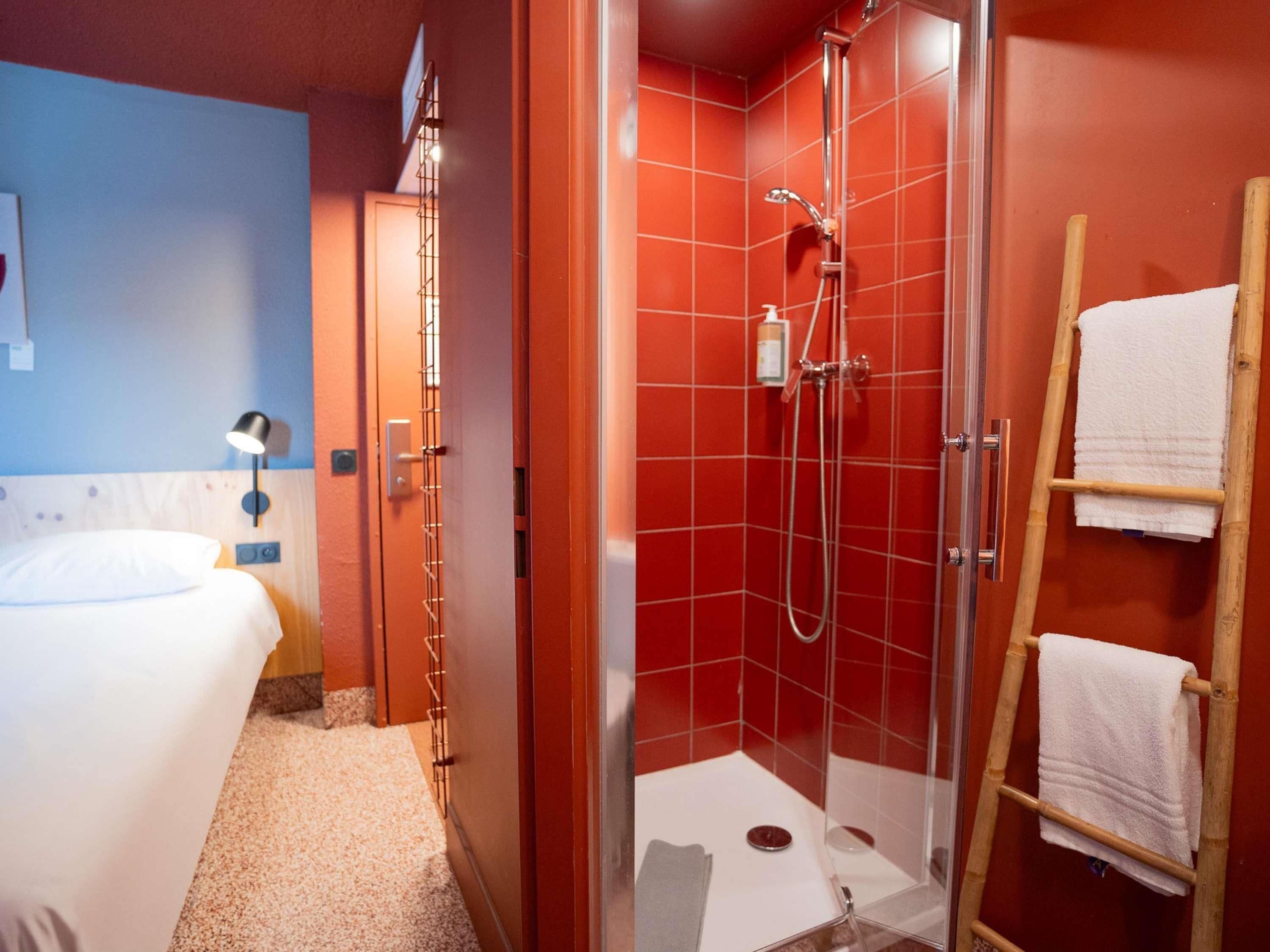 A hotel room with a bed, a shower area with red tiles, and a wooden ladder with towels.