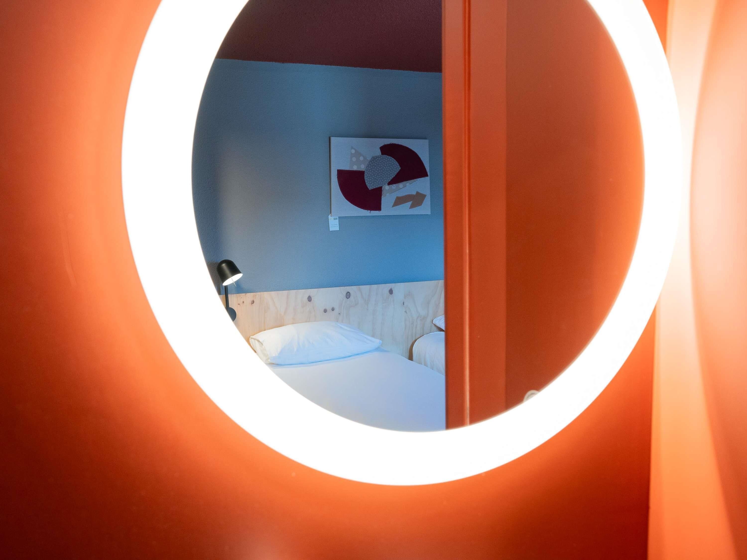 A circular window view of a room with a bed, a wall-mounted lamp, and a piece of abstract art.