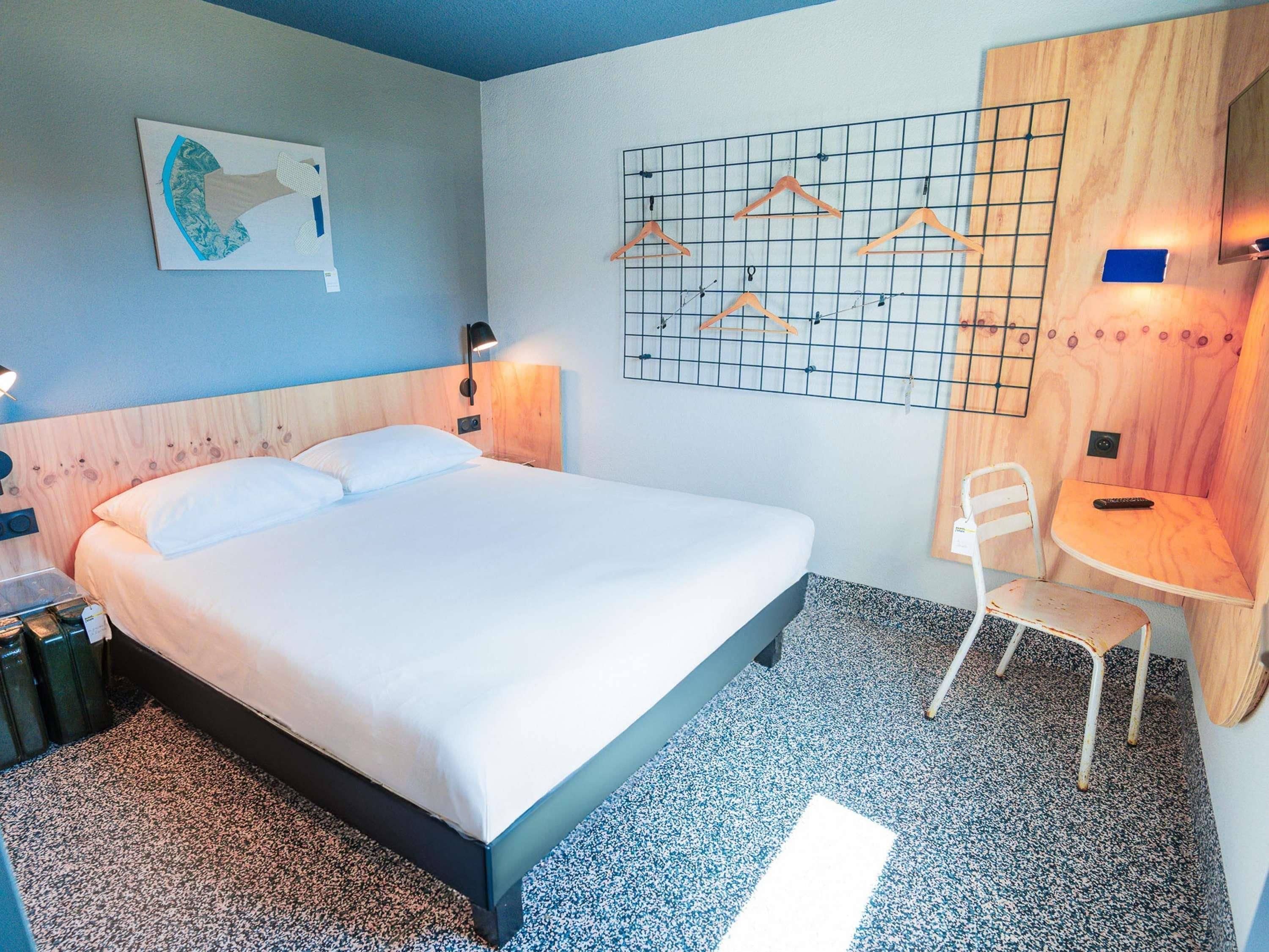 Quarto de hotel com cama, poltrona, escrivaninha, televisão na parede e uma obra de arte na parede.