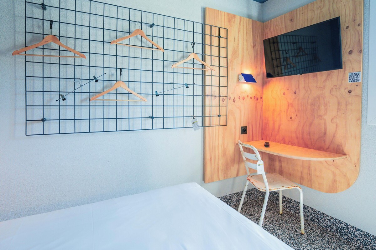 Habitación de hotel compacta con cama, un escritorio pequeño, una silla y un panel en la pared para colgar ropa.