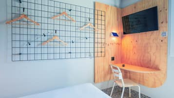 Une chambre d’hôtel de taille réduite, comprenant un lit, un petit bureau, une chaise et un panneau mural servant à suspendre les vêtements.