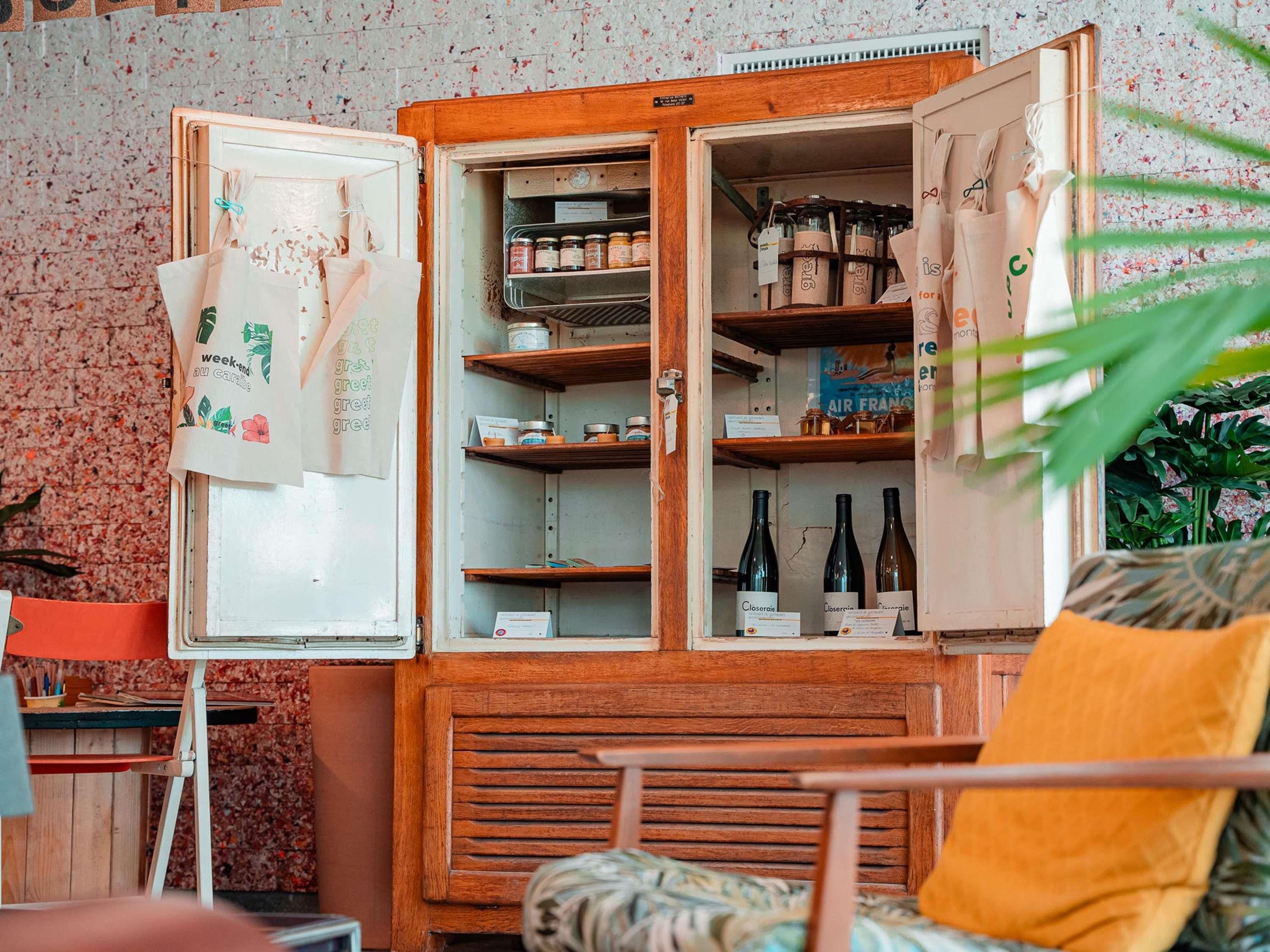 A vintage refrigerator with open shelves containing various items, a chair with a yellow cushion, and a plant in the foreground.