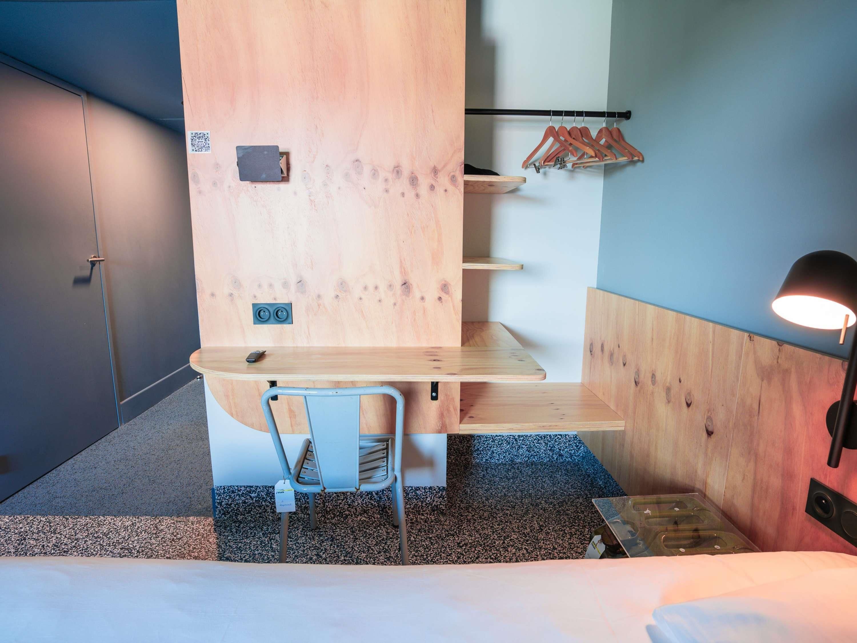 A hotel room with a bed, a desk with a chair, a small shelf, a coat rack, and a wall-mounted lamp.
