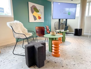 Un interior moderno con una silla verde, una butaca blanca de pelo largo, un cuadro abstracto y colorido, un televisor y una mesa original con taburetes apilados.