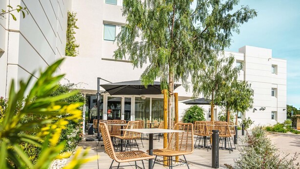 Un patio al aire libre con mobiliario de mimbre, una zona de estar cubierta y un edificio con grandes ventanales al fondo.