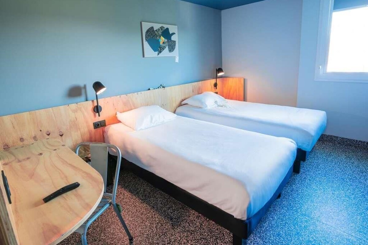 Habitación de hotel con dos camas individuales, cabecero de madera, una mesita redonda y un ventanal.