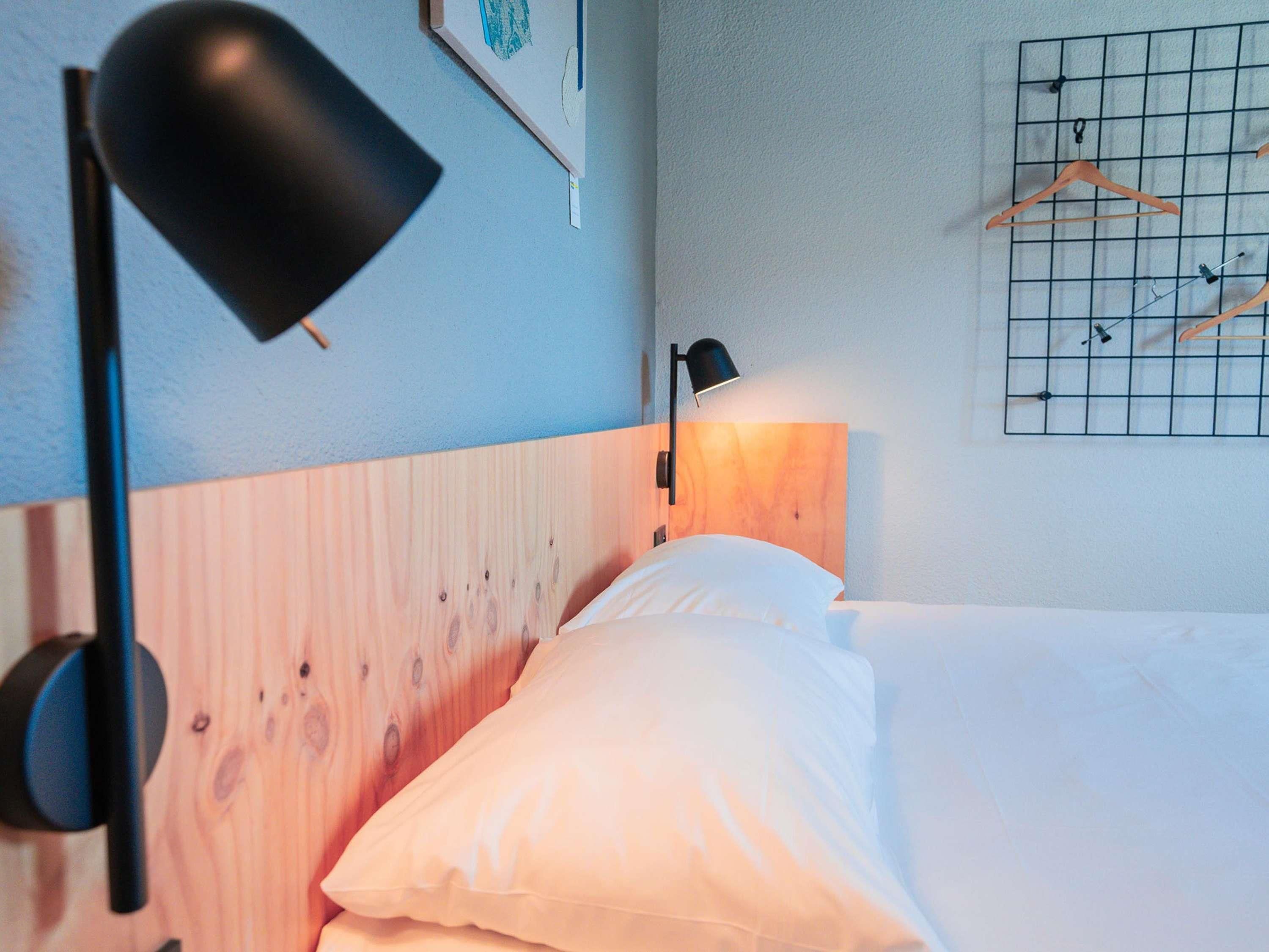 Un dormitorio con cabecera de madera, una cama con ropa de cama blanca, una lámpara de escritorio negra y un aplique de pared.
