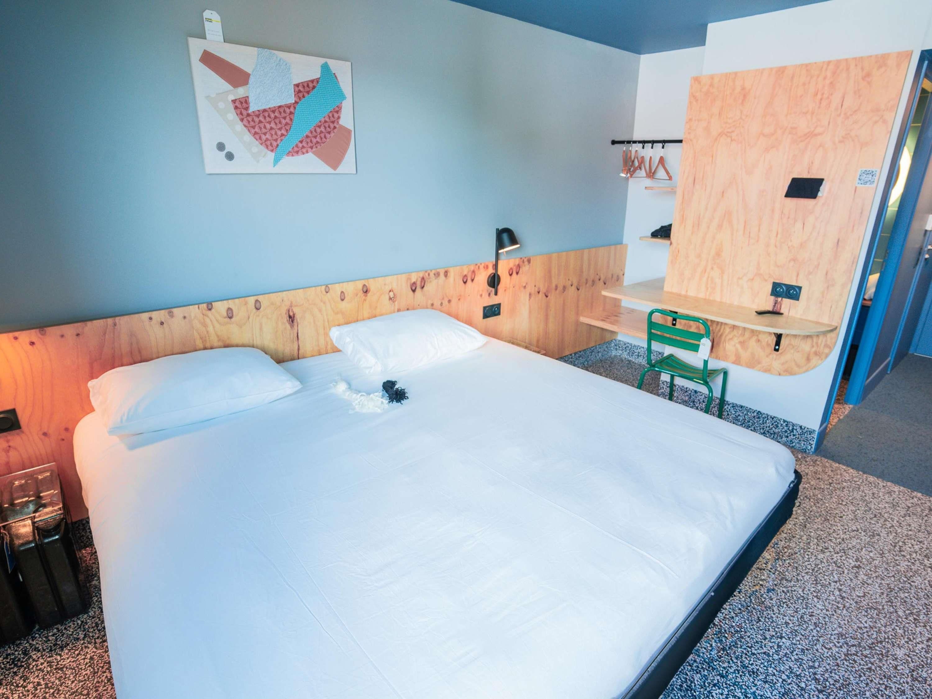 A hotel room with a large bed, a wooden headboard, a small desk with a chair, and a wall-mounted shelf.