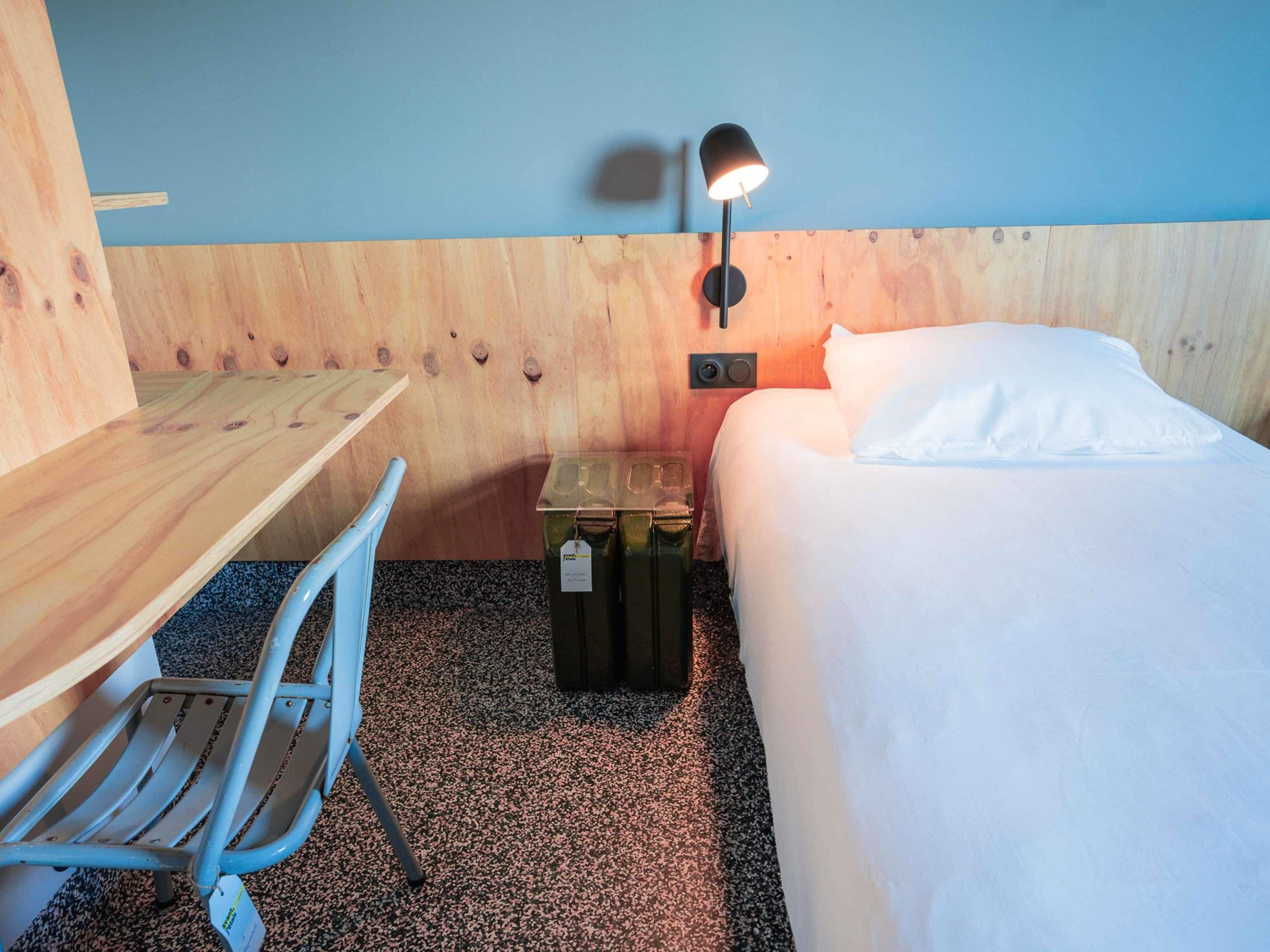 Una habitación con un escritorio de madera, una silla, una cama con ropa blanca, una lámpara de pared y un pequeño cubo de basura.