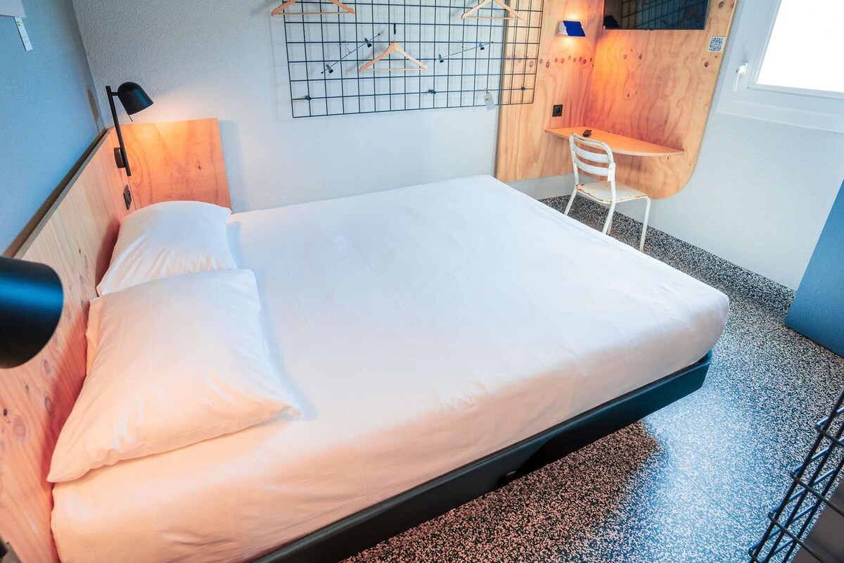 Una cama individual con cabecera de madera, un escritorio con silla y una estructura de rejilla en la pared con perchas de madera.