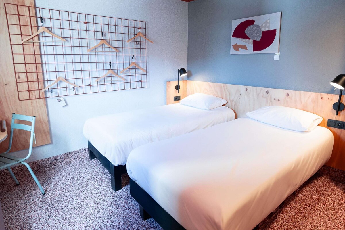 Habitación de hotel con dos camas, cabecero de madera, lámpara de pared y decoración en la pared con diseño de cuadrícula.
