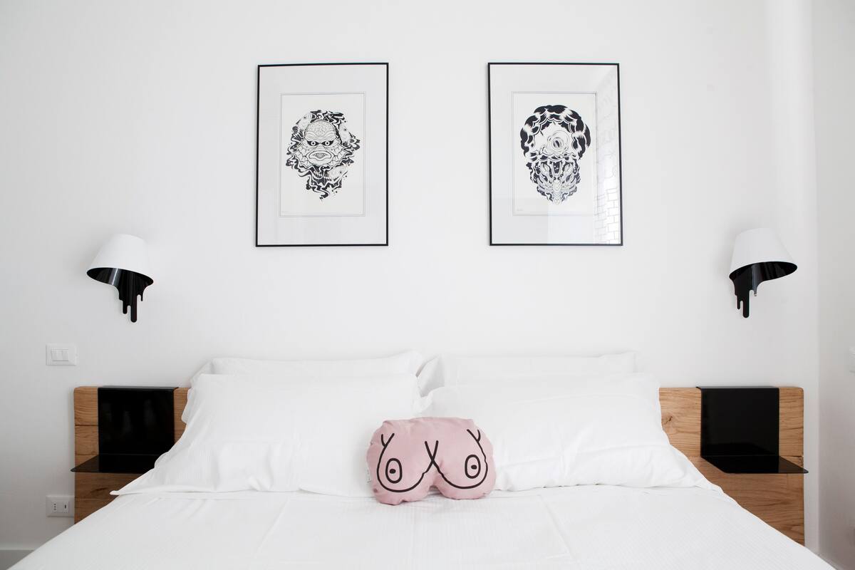 Una cama bien tendida con sábanas blancas, una almohada rosa con un diseño facial y dos cuadros enmarcados sobre la cama.