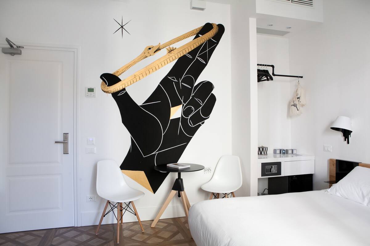 Habitación de hotel con una cama, dos sillas blancas, una mesita redonda, un escritorio con una computadora y una obra de arte en la pared.